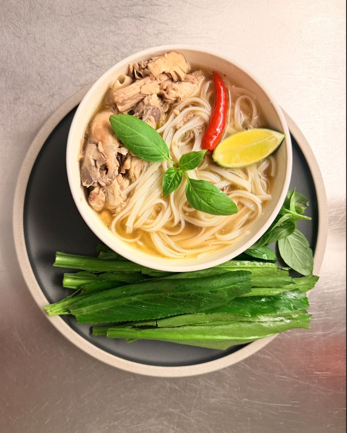 Bowl of chicken noodle soup with basil, chili and lime, and greens on the side