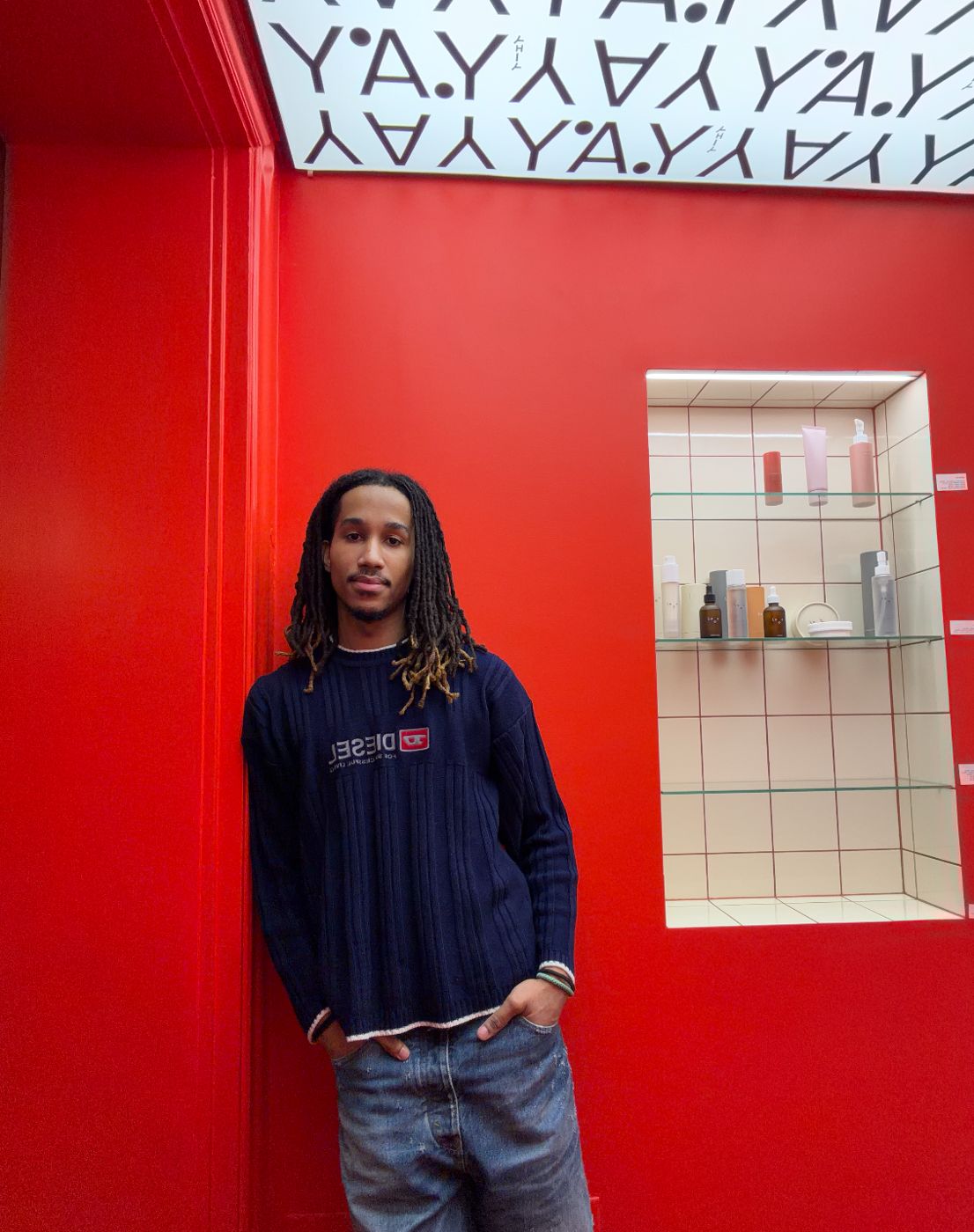 Nabil Nassor leaning against a red wall, hands in pockets, in a navy knit sweater