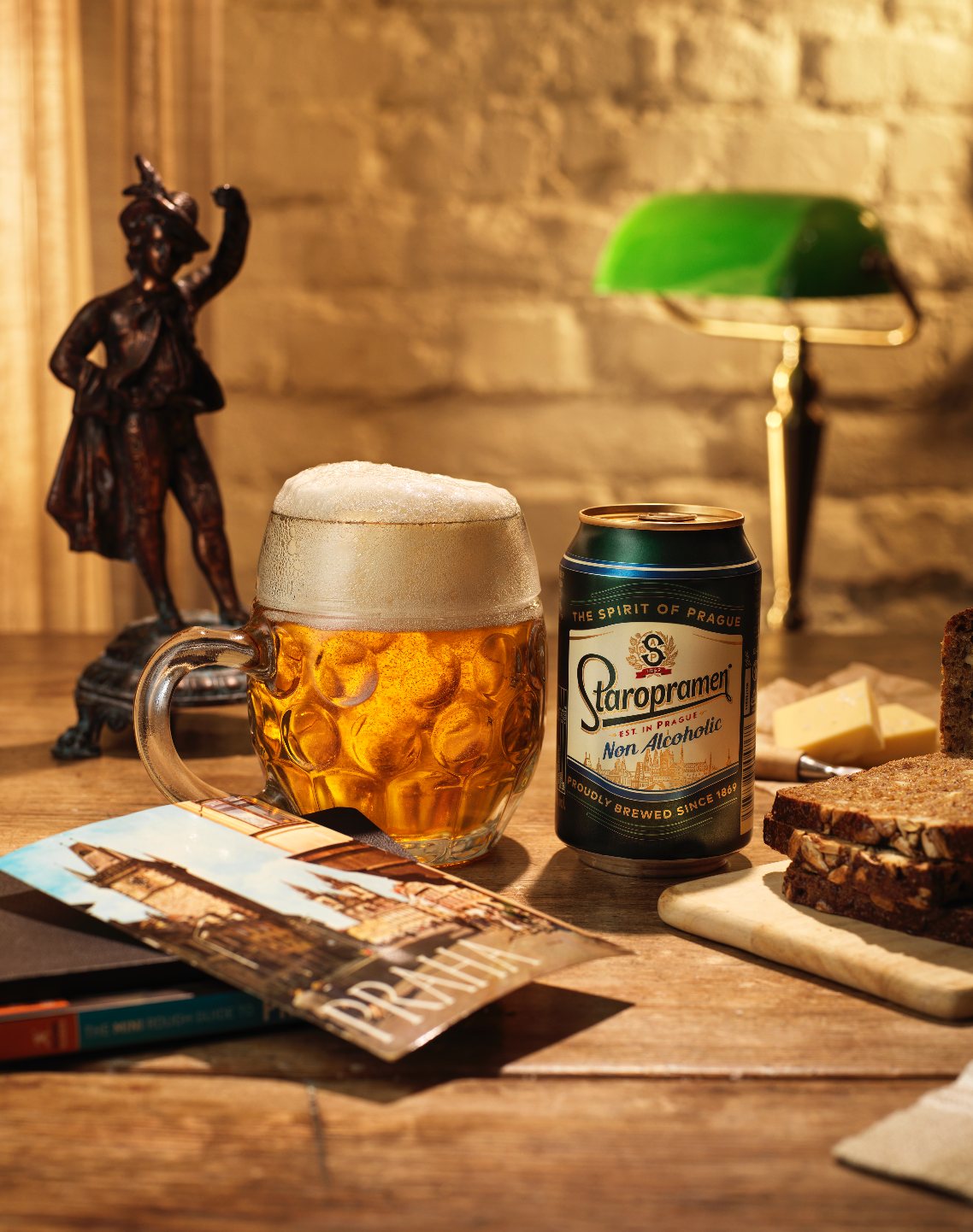 Poster image: a beer mug and a Staropramen can on a wooden table beside a Praha postcard, dark bread and cheese, with a bronze figurine and a green desk lamp behind