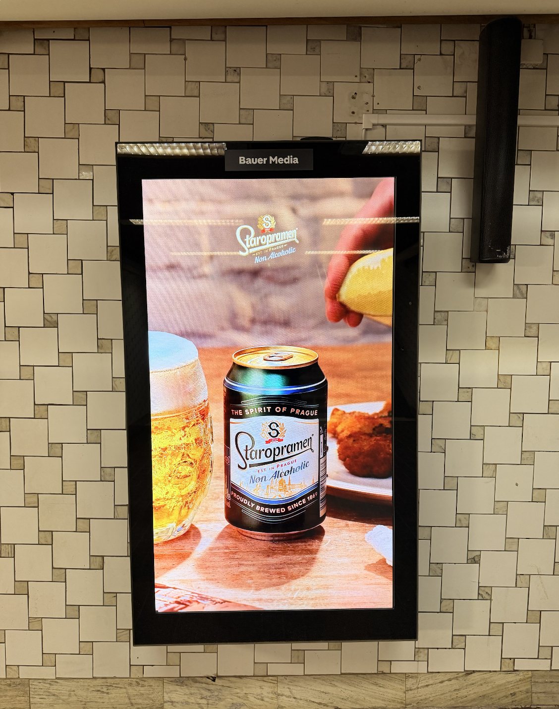 Photo of the Staropramen Non Alcoholic campaign on a Bauer Media digital screen mounted on a tiled wall: a can beside a beer mug and schnitzel, with a hand squeezing a lemon