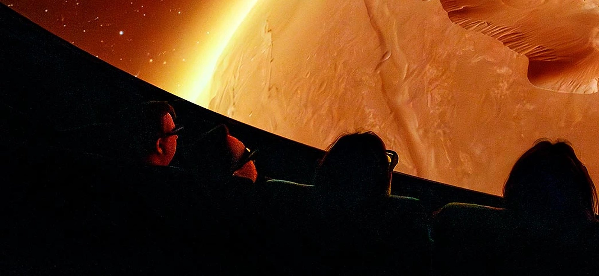 Visitors in dark seats looking up at an orange planet surface projected on the dome